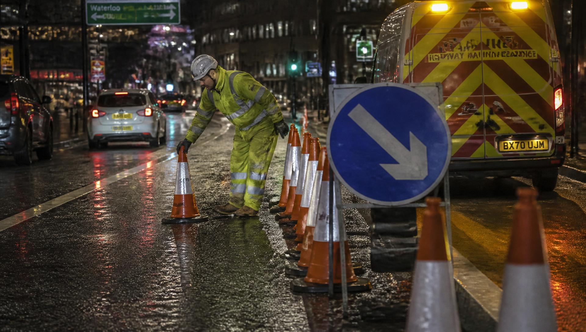 Traffic-management operative placing cones beside a lane closure on a wet London street at night